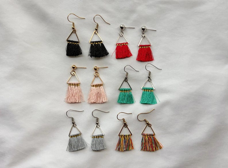 May include: A collection of tassel earrings in various colors, including black, red, pink, green, gray, and multicolored. Each earring features a triangular metal frame and a fringe of thread. The earrings are displayed on a white surface.