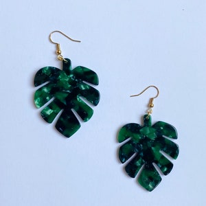 May include: A pair of green and black marbled acrylic earrings in the shape of Monstera leaves. The earrings have gold-colored fish hook ear wires. The earrings are set against a white background.