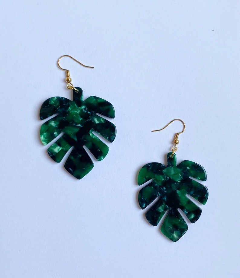 May include: A pair of green and black marbled acrylic earrings in the shape of Monstera leaves. The earrings have gold-colored fish hook ear wires. The earrings are set against a white background.