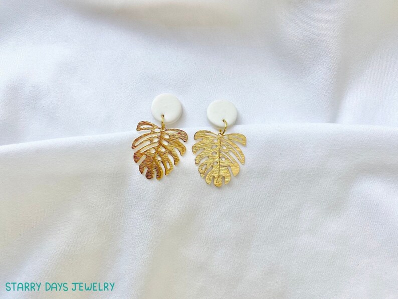 May include: A pair of gold-colored, leaf-shaped earrings with a white circular stud. The earrings are displayed on a white, textured surface. The earrings are from Starry Days Jewelry.