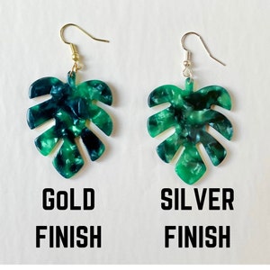 May include: Two leaf-shaped earrings with a marbled green and blue pattern. One earring has a gold-colored hook, and the other has a silver-colored hook. The words "GOLD FINISH" and "SILVER FINISH" are printed below the earrings.
