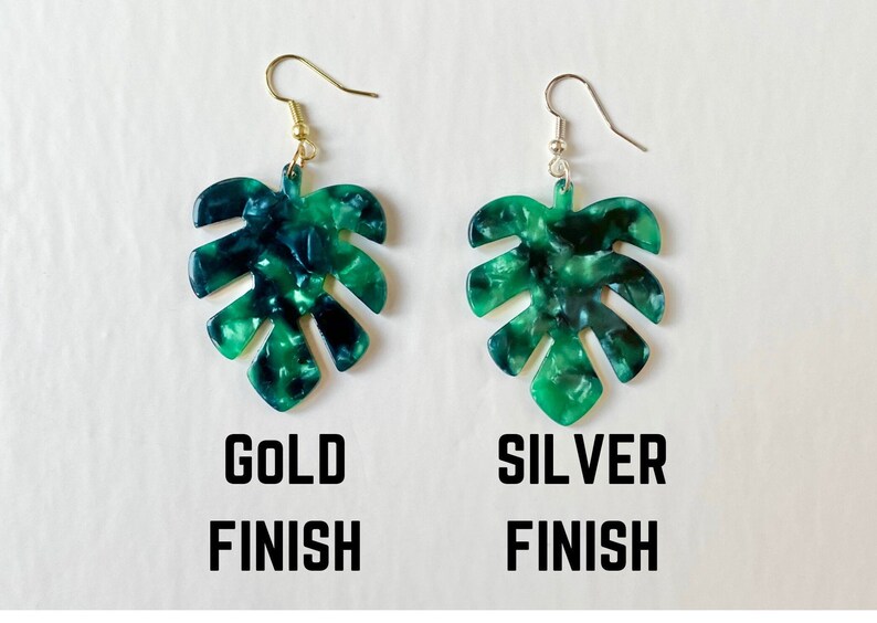 May include: Two leaf-shaped earrings with a marbled green and blue pattern. One earring has a gold-colored hook, and the other has a silver-colored hook. The words "GOLD FINISH" and "SILVER FINISH" are printed below the earrings.