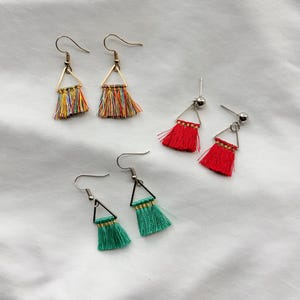 May include: A collection of handmade tassel earrings in various colors. The earrings feature a triangular metal frame with colorful thread tassels. The colors include red, green, and a rainbow mix. The earrings have a hook closure.