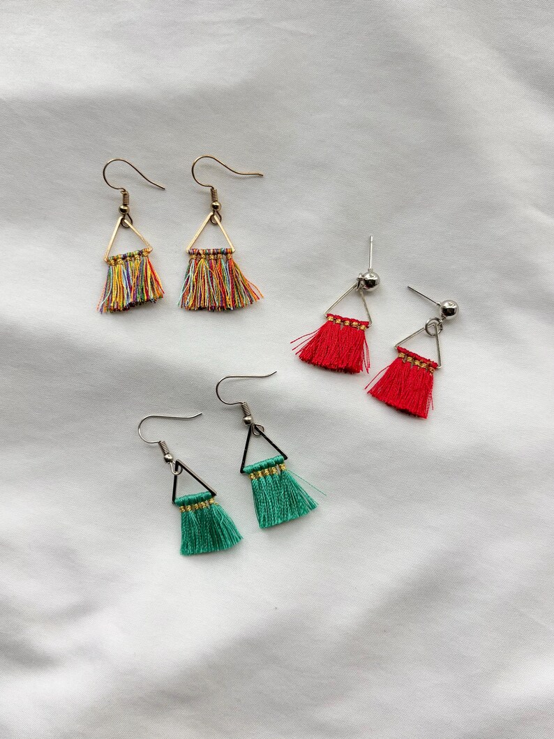 May include: A collection of handmade tassel earrings in various colors. The earrings feature a triangular metal frame with colorful thread tassels. The colors include red, green, and a rainbow mix. The earrings have a hook closure.