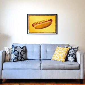 Puede incluir: Un sof&aacute; gris con tres almohadas, dos con un patr&oacute;n azul y blanco y una con un patr&oacute;n geom&eacute;trico amarillo y blanco. Un estampado enmarcado de un hot dog con mostaza y ketchup sobre un fondo amarillo cuelga de la pared sobre el sof&aacute;. Una l&aacute;mpara de pie de tr&iacute;pode negro con una pantalla en forma de flor rosa est&aacute; en la esquina de la habitaci&oacute;n.