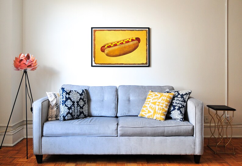 Puede incluir: Un sof&aacute; gris con tres almohadas, dos con un patr&oacute;n azul y blanco y una con un patr&oacute;n geom&eacute;trico amarillo y blanco. Un estampado enmarcado de un hot dog con mostaza y ketchup sobre un fondo amarillo cuelga de la pared sobre el sof&aacute;. Una l&aacute;mpara de pie de tr&iacute;pode negro con una pantalla en forma de flor rosa est&aacute; en la esquina de la habitaci&oacute;n.