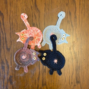 May include: Four crocheted cat-shaped coasters in different colors: pink, gray, brown, and black. Each coaster has two large button eyes and a small button nose.