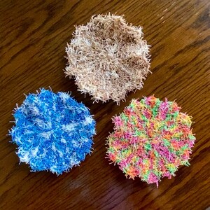 Puede incluir: Tres paños de cocina de ganchillo en diferentes colores. Uno es marrón, otro es azul y blanco, y el último es una mezcla de rosa, verde, naranja y amarillo.