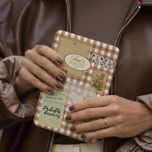Könnte beinhalten: Ein rechteckiges Etui mit braun-weißem Vichy-Muster. Das Etui hat dekorative Etiketten mit den Texten "Soul Mates", "Kinole Girly" und "Probably Reading". Das Etui wird vor einer braunen Lederjacke gehalten.