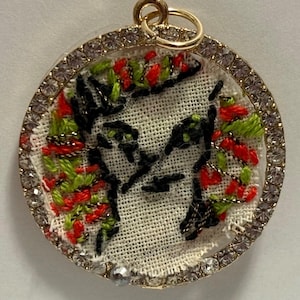 May include: A circular pendant featuring a piece of white fabric embroidered with a black outline of a face, accented by red and green floral stitching. The pendant is framed by a gold-toned metal border encrusted with clear rhinestones.