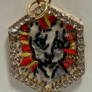 May include: A hexagonal pendant featuring a central piece of off-white fabric embroidered with black, grey, yellow, and red thread. The embroidery is framed by a border of small, clear, sparkling rhinestones set in a gold-toned metal frame.