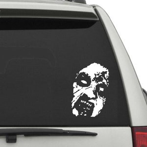 May include: A white vinyl sticker of a scary, distorted face with a wide open mouth and sharp teeth. The sticker is on the back window of a silver car.