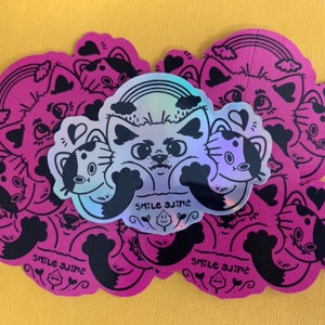 Op de afbeelding: Een set van 10 roze en zwarte stickers met een kat met een regenboog en de tekst "Smile Slime".