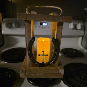 May include: Rustic wooden lantern with a horseshoe and cross design. The lantern features a faux candle with a warm, inviting glow. A jute rope handle adds a touch of country charm. The lantern is a decorative piece.