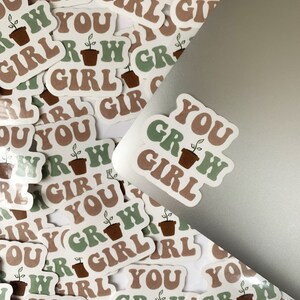 Puede incluir: Primer plano de varias pegatinas con el texto "You Grow Girl" en una fuente retro. Las pegatinas muestran una planta verde en una maceta marrón. Las pegatinas están en un portátil plateado.