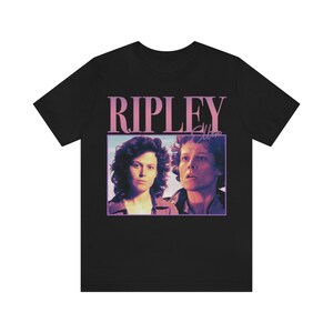 May include: Black t-shirt with a pink graphic of Ripley from the movie Alien. The graphic includes a portrait of Ripley and the text "RIPLEY" in pink.