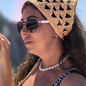 Pode incluir: Um lenço de crochê bege com um padrão triangular vazado, combinado com óculos de sol aviador escuros, um colar de pérolas brancas e uma regata preta com o texto branco CALVIN KLEIN nas alças.