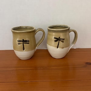 May include: Two ceramic mugs featuring a two-tone design with a tan upper section and a white lower section. Each mug is decorated with a dark brown, hand-painted dragonfly motif on the front and includes a curved handle on the side.