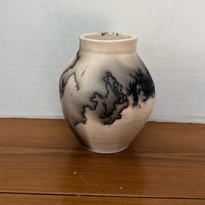 May include: A small, rounded ceramic vase with a matte, off-white surface featuring dark, smoky black horsehair markings that create irregular, flowing lines and patterns across the exterior of the vessel, which has a narrow, open neck.