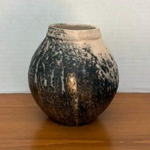 May include: A round ceramic vase with a narrow neck and a wide body. The surface features a textured finish with a gradient of cream at the top transitioning into a dark, mottled black and brown pattern that covers the lower portion of the vessel.