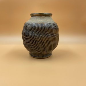 Puede incluir: Un pequeño jarrón de cerámica marrón con un diseño texturizado ondulado. El jarrón tiene un borde marrón claro y una base ligeramente más ancha.
