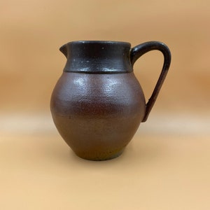 May include: A brown ceramic pitcher with a dark brown rim and a handle. The pitcher has a textured surface.