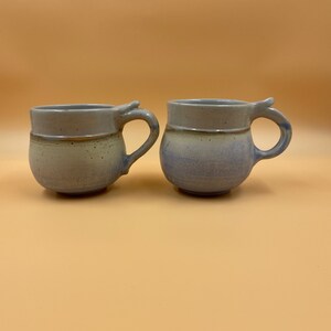 Puede incluir: Dos tazas de cerámica con un esmalte azul claro y beige. Las tazas tienen un diseño de asa único.