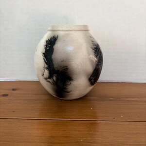 May include: A round, off-white ceramic vase with a smooth, matte finish. The surface features dark, wispy, black smoke-like markings that wrap around the sides. The vase has a narrow, slightly flared rim and a rounded body that tapers at the base.