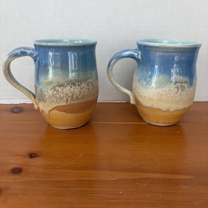 May include: Two ceramic mugs featuring a glossy glaze with a blue top section that transitions into a speckled white middle and a solid tan base. Each mug has a curved handle and a rounded body, showcasing a layered color effect.