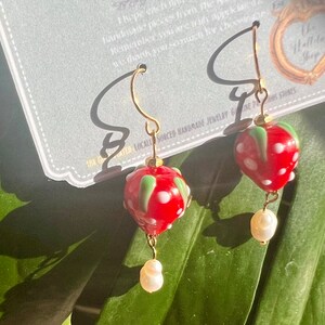 May include: A pair of gold dangle earrings with red and white glass strawberry charms and white pearl accents. The earrings are displayed on a white card with the text "Locally Sourced Handmade Jewelry & Gift Stones" and "The Hellola Shop".