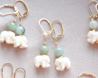 May include: Gold-tone leverback earrings featuring a carved ivory-colored elephant charm and a pale green bead. The earrings are displayed against a neutral background, showcasing their delicate design.