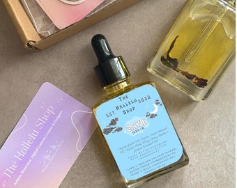 Può includere: Un piccolo flacone di vetro con un contagocce nero in cima, riempito di un liquido giallo chiaro. Il flacone ha un'etichetta blu con testo bianco che recita "THE EST. HALLELU 2020 SHOP CLOUD BODY OIL" e una descrizione degli ingredienti. Il flacone è su una superficie blu chiaro.