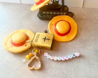May include: A collection of novelty items including miniature straw hats with red bands, a gold-colored Holy Bible, and a keychain with letter beads spelling out a word. The hats are yellow and red. The keychain has a gold heart-shaped clasp.