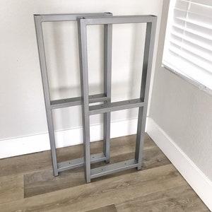 May include: Two silver metal table legs with a rectangular frame design. The legs are standing on a wood floor.