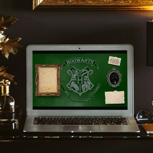May include: A silver laptop computer with a green screen displaying the Hogwarts crest and the text "Hogwarts School of Witchcraft and Wizardry." The laptop is on a dark surface with a gold picture frame, a gold lamp, and a gold and black decorative object.