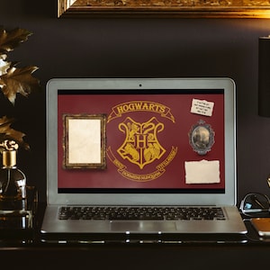May include: A laptop computer with a red background showing the Hogwarts crest and the words "Hogwarts School of Witchcraft and Wizardry". The laptop is on a dark surface with a gold picture frame, a small piece of paper, and a gold lamp.
