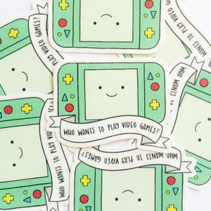 May include: A collection of stickers featuring a cartoon-style handheld video game console in shades of green. Each console has a smiling face and the text "WHO WANTS TO PLAY VIDEO GAMES?" on a banner.