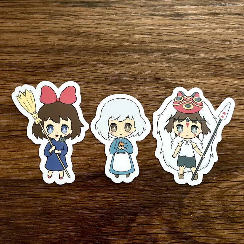 May include: Three anime-style stickers featuring characters with large eyes. The first sticker shows a girl in a blue dress with a red bow, holding a broom and a black cat. The second sticker is a girl in a blue dress and white apron. The third sticker is a girl with a mask and spear.