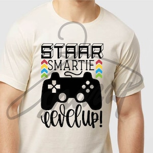 Puede incluir: Camiseta color crema con un gráfico negro de un mando de videojuegos y las palabras "STAAR SMARTIE level up!" en negro. El texto está en una fuente lúdica, con acentos de flechas de colores.