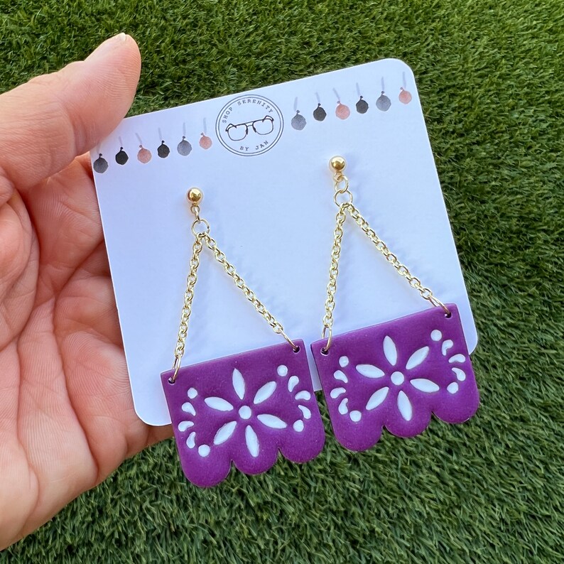 May include: Purple earrings with a floral design and gold-coloured chain. The earrings are rectangular with a scalloped bottom edge and a white floral pattern. The earrings are displayed on a white card with the text "Shop Serenity by Jam".
