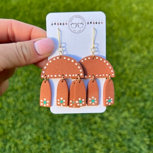 May include: Terracotta and white clay earrings with a geometric design. The earrings feature a half-circle shape with white dots and three teardrop shapes with white dots and a turquoise accent.