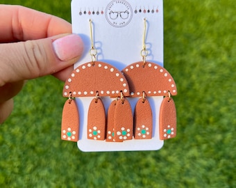 May include: Terracotta and white clay earrings with a geometric design. The earrings feature a half-circle shape with white dots and three teardrop shapes with white dots and a turquoise accent.