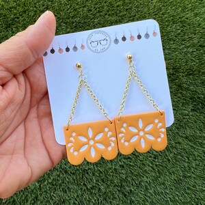May include: Orange and white floral design earrings with gold-coloured chain and stud attachments. The earrings are displayed on a white card with the shop's logo. The earrings have a scalloped edge.