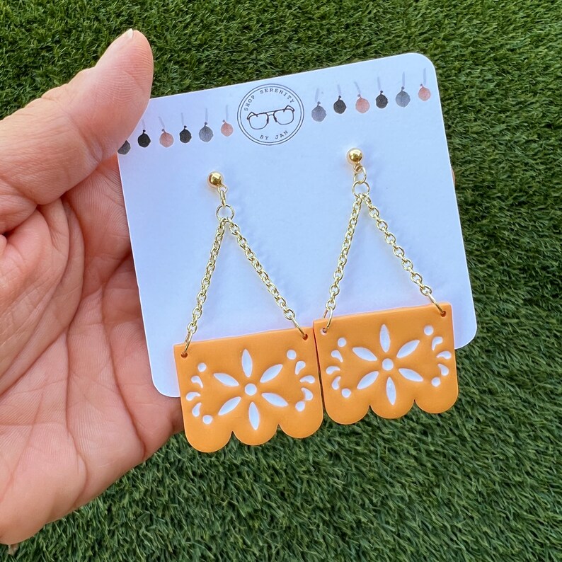 May include: Orange and white floral design earrings with gold-coloured chain and stud attachments. The earrings are displayed on a white card with the shop's logo. The earrings have a scalloped edge.