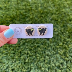 K&ouml;nnte beinhalten: Ein Paar schwarze Katzen-Ohrstecker mit gr&uuml;nen Augen auf beigefarbenem Hintergrund. Die Ohrringe befinden sich auf einer Karte mit dem Text "Shop Serenity by Jan" und einer Website-Adresse.