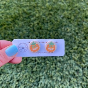 K&ouml;nnte beinhalten: Ein Paar K&uuml;rbis-Ohrringe mit einem gr&uuml;nen Stiel und einem Jack-o'-Lantern-Gesicht. Die Ohrringe befinden sich auf einer wei&szlig;en Karte mit dem Text "shop serenity by jan" und einer Website-Adresse.