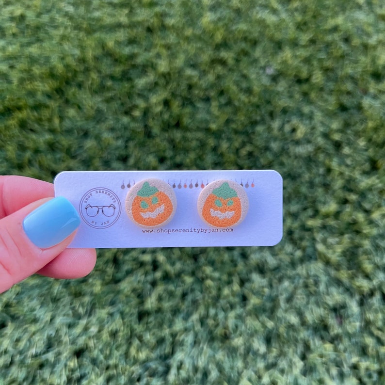 K&ouml;nnte beinhalten: Ein Paar K&uuml;rbis-Ohrringe mit einem gr&uuml;nen Stiel und einem Jack-o'-Lantern-Gesicht. Die Ohrringe befinden sich auf einer wei&szlig;en Karte mit dem Text "shop serenity by jan" und einer Website-Adresse.