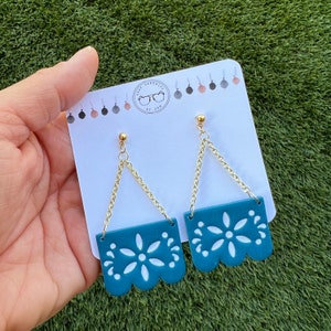 May include: Teal dangle earrings with a floral cut-out design and gold-coloured chain. The earrings are attached to a white card with the text "Shop Serenity by Jan" and a logo. The earrings are displayed on a green surface.