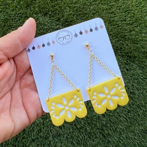 May include: Yellow earrings with a floral design and gold-coloured chain. The earrings are displayed against a white card with the text "Shop Serenity by Jam". The earrings have a scalloped edge.