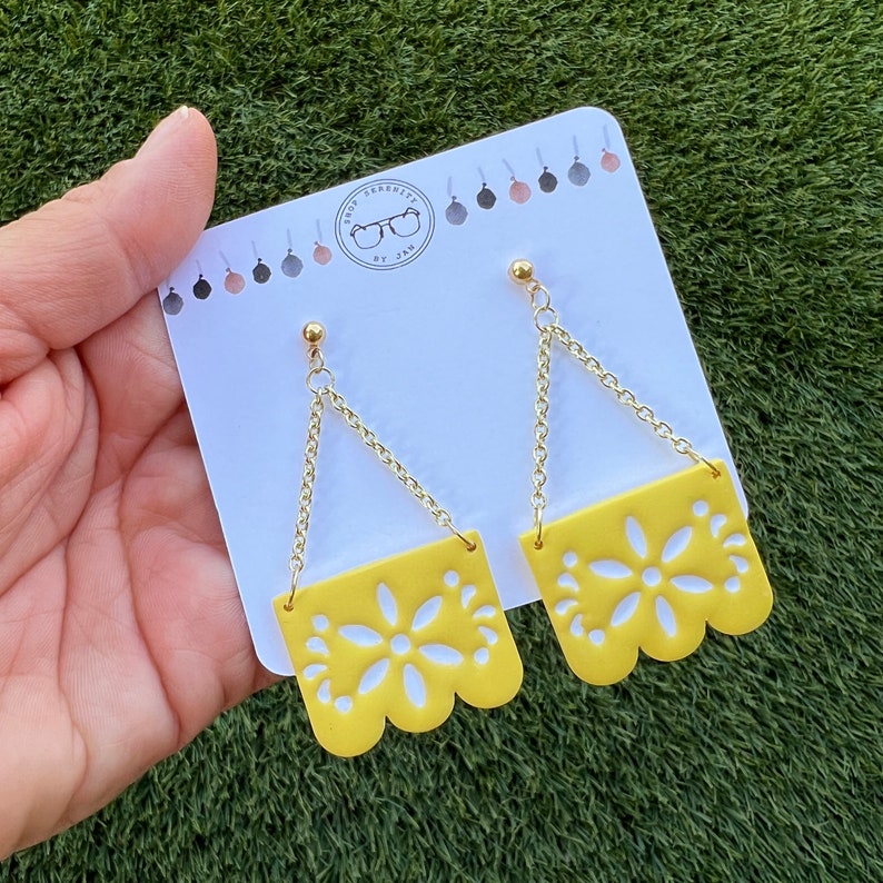 May include: Yellow earrings with a floral design and gold-coloured chain. The earrings are displayed against a white card with the text "Shop Serenity by Jam". The earrings have a scalloped edge.